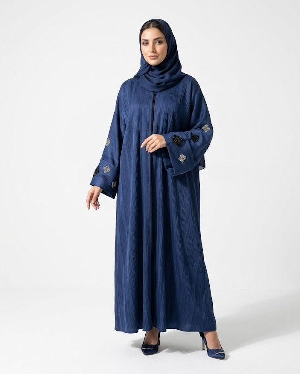 A woman modeling a long navy blue abaya with matching hijab and navy pointed shoes against a light gray studio background.